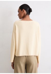 Helle beigefarbene Strickjacke mit weitem Schnitt, breitem Ausschnitt und langen Ärmeln. Weicher Stoff, leicht verkürzte Länge, kombiniert mit olivgrünen Hosen.