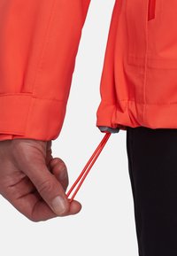 Veste orange vif avec une finition texturée ; main ajustant le cordon élastique à la taille ; tissu noir contrastant en dessous.