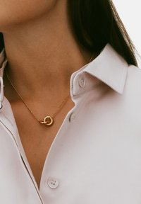 Gold chain necklace featuring interlocking circular links, worn over a white collared shirt with buttons. Smooth texture, minimal design.