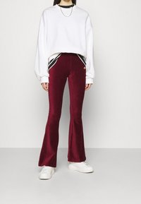 Burgundy utsvängda sammetbyxor i kombination med en vit oversized sweatshirt och vita sneakers. Har en kontrasterande svart rand och logotypdetalj.