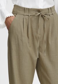 Khaki plisované nohavice s pásom na stiahnutie, s detailom gombíka a hladkou textúrou. Predné vrecká viditeľné na každej strane.