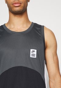 Homme portant un haut de sport sans manches Nike Force gris foncé et noir avec un écusson logo sur la poitrine devant un fond uni.