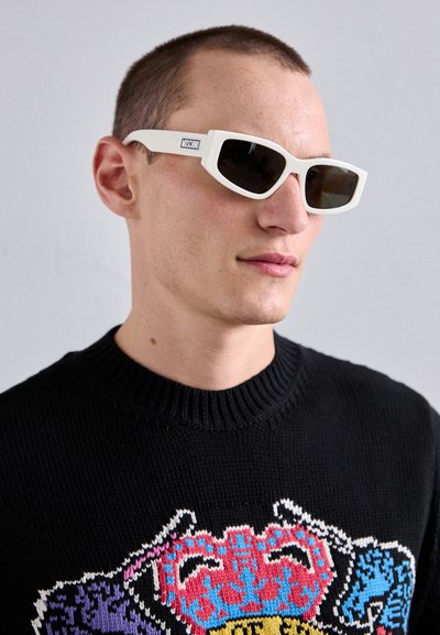 Bele angularne sončna očala s temnimi lečami. Mat zaključek. Model nosi črn pulover z barvitim grafičnim vzorcem.