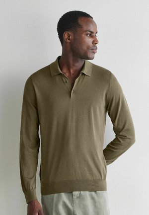 Mężczyzna ubrany w oliwkowo zieloną koszulkę polo z długim rękawem, patrzący w bok na jednolitym jasnym tle.