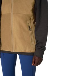 Person iført en beige fleecejakke med lynlås sidelomme, mørke lange ærmer og blå leggings, hånden afslappet ved siden af kroppen.