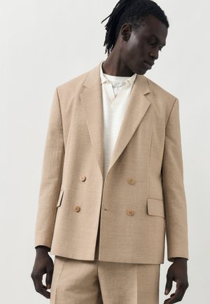 Sort mand med dreadlocks iført en beige dobbeltradet dragt og hvid skjorte med krave, der ser nedad og mod sin venstre side mod en lys baggrund.