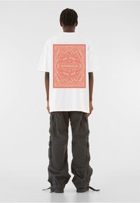White oversized t-shirt featuring a red graphic pattern on the back with the text "FREE SPIRIT" and "M.GONZALES." Paired with black cargo pants.