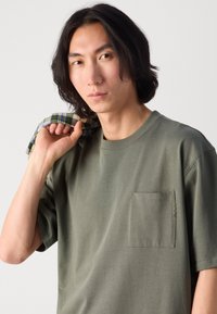 T-shirt en coton vert olive avec une poche poitrine, des manches courtes et des détails de couture visibles. Le modèle tient une chemise à carreaux sur son épaule.