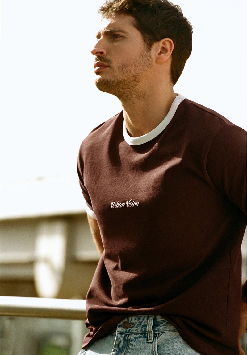 Jeune homme aux cheveux courts bouclés portant un t-shirt bordeaux foncé "Urban Vision" et un jean bleu clair, debout à l'extérieur près d'une rambarde.