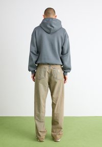 Grauer Kapuzenpullover mit entspannter Passform, kombiniert mit beige Baggy-Jeans mit zwei Gesäßtaschen und einem Logo-Patch, von hinten betrachtet.