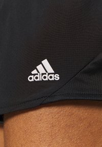 Sorte atletikshorts med hvidt Adidas-logo på kanten mod synlig hud på en persons lår.