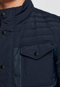 Námořnická prošívaná bunda s vysokým límcem, černými patentkami a klopovou kapsou na prsou s hladkým povrchem.