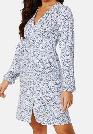 Femme portant une robe blanche mi-genou avec imprimé floral bleu, manches longues, devant boutonné, et décolleté en V.