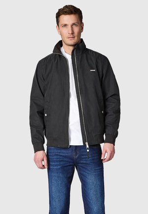 Mężczyzna stoi, ubrany w czarną kurtkę zapinaną na zamek na białą koszulę i niebieskie dżinsy na jednolitym szarym tle.