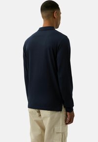 Marineblaues Langarm-Polohemd mit klassischem Kragen, weicher Textur und seitlichen Schlitzen; kombiniert mit hellbeigen Cargo-Hosen.