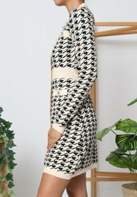 Femme portant une robe cardigan à manches longues avec un motif pied de poule noir et blanc, se tenant à l'intérieur près de plantes d'intérieur vertes et d'un cadre en bois.