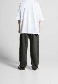 Persoon in een losse witte korte mouwen shirt, donkergrijze wijde broek en witte schoenen die op een effen grijze vloer staat.