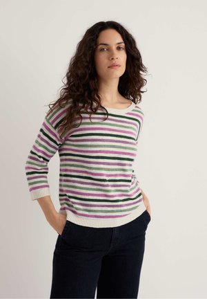 Frau mit langen lockigen dunklen Haaren, die einen rosa, grünen und weißen gestreiften Pullover und schwarze Hosen trägt, steht mit den Händen in den Taschen.