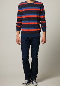 Sweter w niebiesko-czerwone paski z okrągłym dekoltem, zestawiony z ciemnoniebieskimi dżinsami i białymi tenisówkami. Materiał wydaje się miękki, a krój dopasowany.