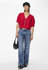 Top rosso corto con maniche a palloncino e nodo anteriore, abbinato a jeans in denim blu a vestibilità ampia. Gli accessori includono una piccola borsa nera.