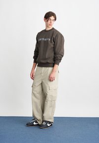 Sweatshirt maro cu guler rotund, având un logo albastru deschis, asortat cu pantaloni cargo bej care includ buzunare laterale, și adidași negri cu accente albe.