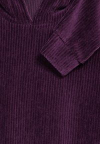 Tessuto di velluto a coste viola con texture a coste verticali. Presenta una scollatura a V e maniche lunghe con polsini a coste. Aspetto morbido e peluche.