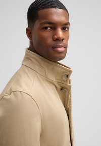 Beige jas met een hoge kraag en knoopdetails, met een gladde textuur. Het gezicht van het model is gedeeltelijk zichtbaar tegen een neutrale achtergrond.