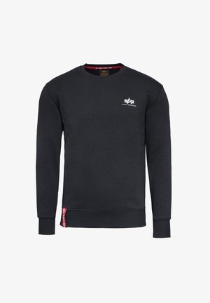 Sweat-shirt noir à col rond avec le logo Alpha Industries sur la poitrine et une étiquette rouge près de la ceinture sur le côté avant gauche.