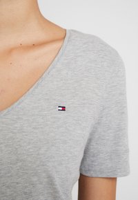 Maglietta grigia a V in tessuto morbido con maniche corte, caratterizzata da un piccolo logo ricamato in blu, rosso e bianco sopra il lato sinistro del petto.