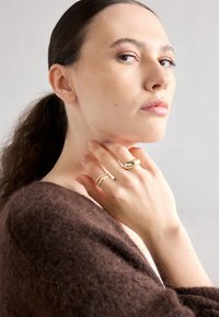 Aukso žiedai ant pirštų su lygiu ir tekstūruotu paviršiumi. Modelis vilki rudą, švelnų drabužį. Paveikslas išryškėja prieš šviesiai pilką foną.
