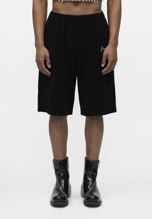 Person som har på seg svarte knelang shorts med broderi og skinnende svarte ankelstøvler med front glidelåser, stående mot en hvit bakgrunn.