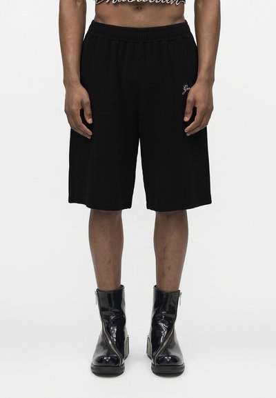Personne portant un short noir longueur genou avec broderie et des bottines noires brillantes à fermeture éclair frontale, debout sur fond blanc.