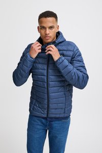 Jachetă albastră căptușită cu glugă detașabilă, model cu quilt vertical și fermoar frontal. Purtată cu blugi.