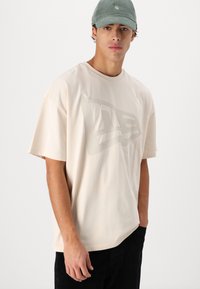 Gaiši bež oversize T-krekls no kokvilnas, ar lielu, tonālu zīmola logo uz priekšpuses. Modelis pozē ar zaļu cepuri un tumšām biksēm.