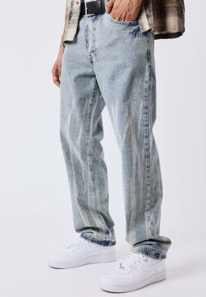 Man draagt acid-wash jeans met een zwarte riem, een geruit overhemd en witte Nike sneakers, staat met één hand in zijn zak.