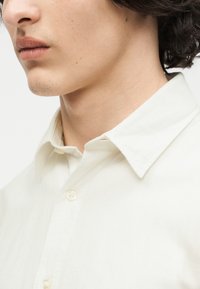 Νεαρό άτομο με ελαφρώς σγουρά σκουρόχρωμα μαλλιά, που φοράει εκρού πουκάμισο με γιακά και κουμπιά, εμφανίζεται από τον λαιμό έως το πηγούνι.