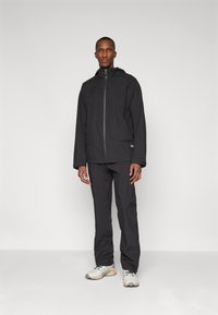 Veste noire imperméable avec capuche et fermeture éclair, associée à un pantalon noir assorti. Le mannequin se tient sur un fond neutre, portant des chaussures de couleur claire.