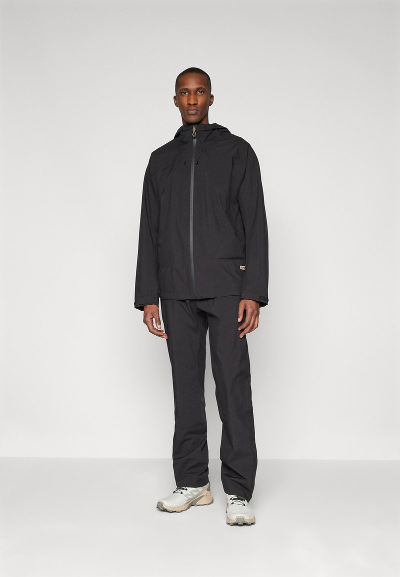Veste noire imperméable avec capuche et fermeture éclair, associée à un pantalon noir assorti. Le mannequin se tient sur un fond neutre, portant des chaussures de couleur claire.