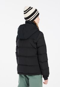 Doudoune noire avec un design matelassé et une capuche. Portée avec un bonnet en tricot crème et noir à rayures. Les détails incluent des poignets côtelés.
