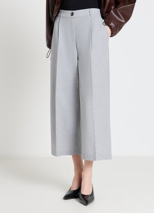 Donna che indossa pantaloni larghi grigio chiaro tagliati sopra la caviglia con pieghe frontali, abbinati a tacchi neri a punta e una giacca marrone.