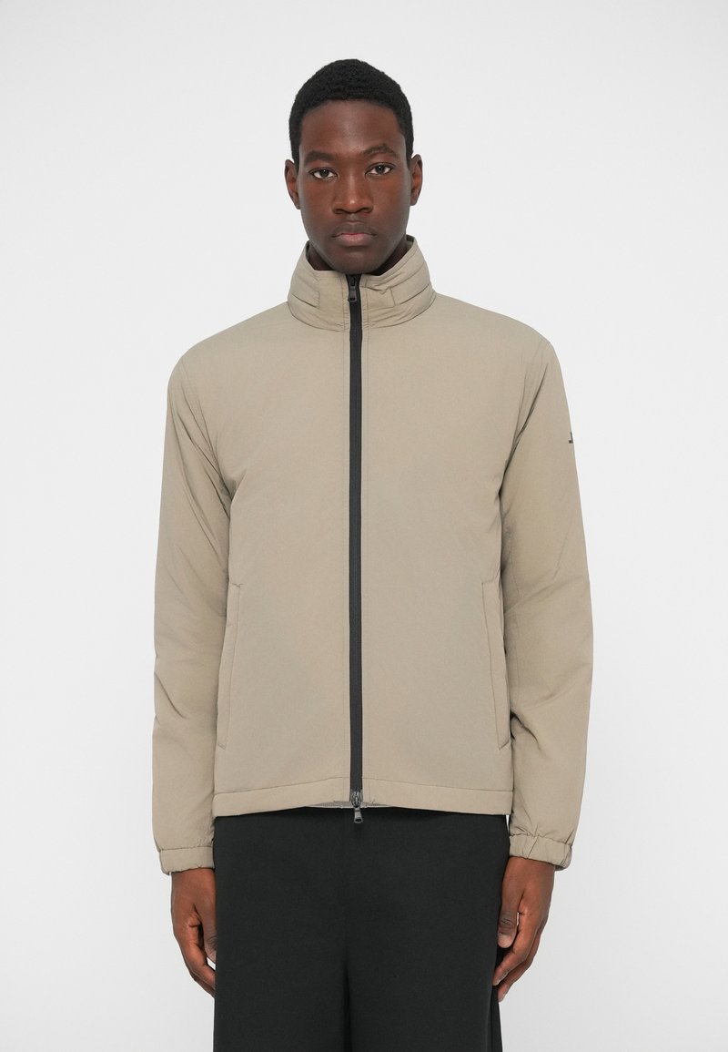 Jachetă ușoară de culoare bej, cu guler înalt, fermoar frontal, două buzunare laterale și manșete elastice. Material neted cu un finisaj mat.