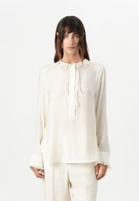 Lettvekts, kremfarget bluse med knyting i halsen, frynset kant og lange ermer. Stoffet har en jevn tekstur og subtil glans.