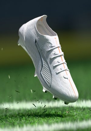 White football cleat with textured upper, orange accents, laces, and spiked sole, suspended above green turf, with grass blades disturbed below.