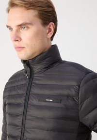 Giacca a vento nera con colletto alto, cuciture orizzontali e patch con logo sul petto. Il materiale sembra lucido e liscio.