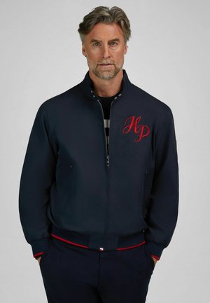 Mittelalter Mann mit grauem Haar und Bart, trägt eine marineblaue Reißverschlussjacke mit roten "HP"-Initialen und hat die Hände in den Taschen.