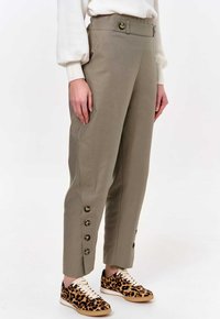 Light khaki trousers with a fitted waist, featuring five gold buttons along the side. Paired with leopard print sneakers.