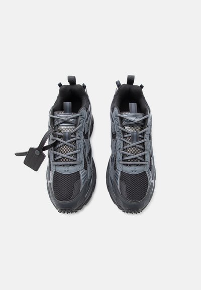 Ζευγάρι αθλητικά παπούτσια σε μαύρο και γκρι χρώμα με επενδύσεις από δίχτυ και συνθετικά υλικά, δεμένα με κορδόνια, το ένα παπούτσι έχει μαύρη ετικέτα προσαρτημένη στα κορδόνια.