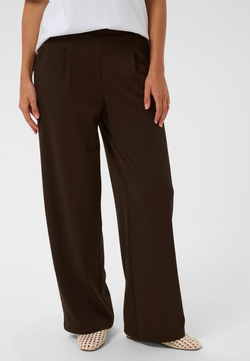 Personne portant un pantalon large marron foncé, une chemise blanche et des chaussures blanches tissées sans lacets, debout devant un fond clair uni.