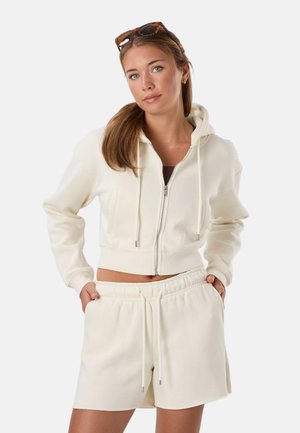 Giovane donna che indossa una felpa con zip color crema e pantaloncini abbinati con coulisse, con occhiali da sole appoggiati sulla testa, posa con le mani nelle tasche.