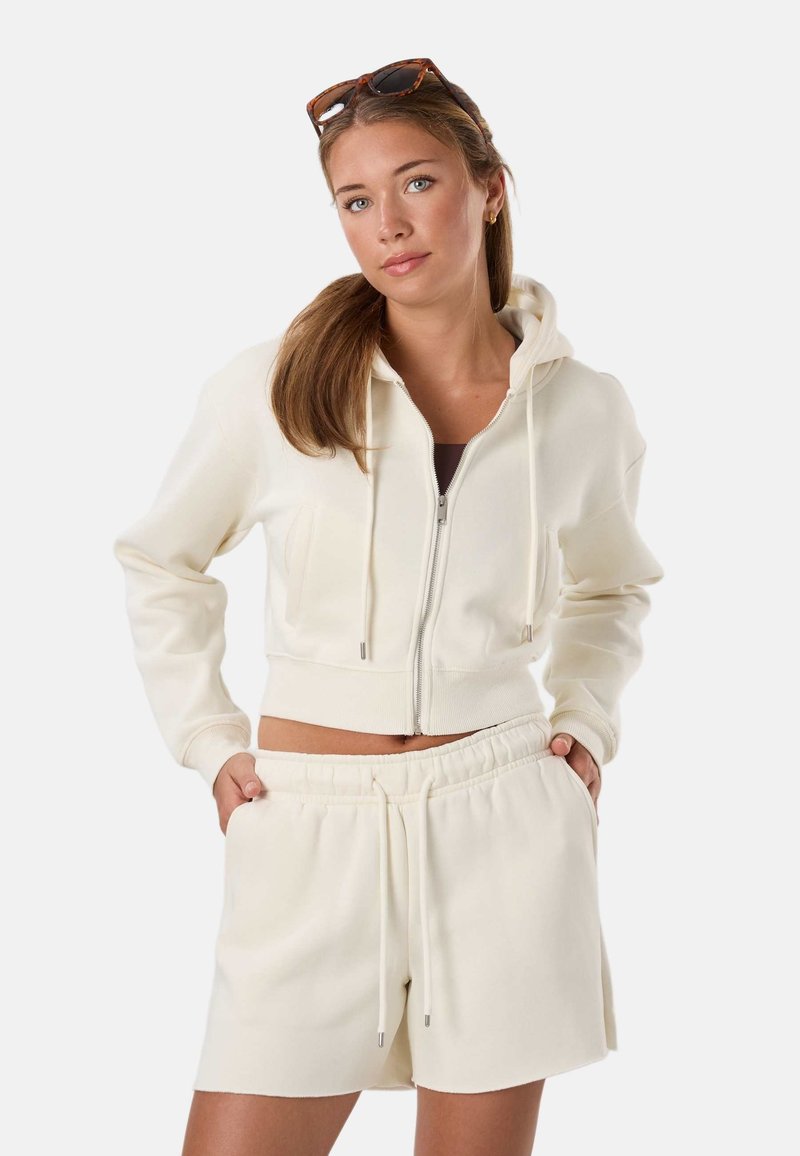 Mujer joven con sudadera con capucha color crema con cremallera y pantalones cortos a juego con cordón, con gafas de sol sobre la cabeza, posando con las manos en los bolsillos.